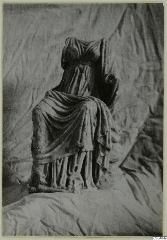 Headless seated draped statue in marble, photographed against a crumpled cloth backdrop.