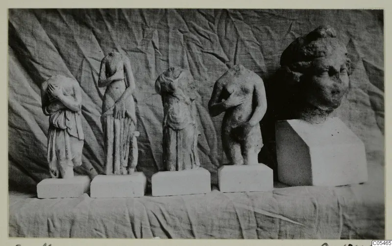 Four small standing stone figures on white plinths and a larger carved head on a block, photographed against draped cloth.