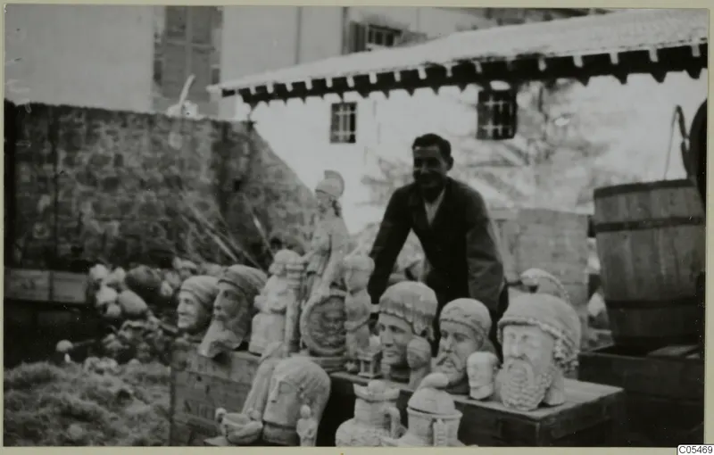Smiling man leaning over a crate of carved stone heads and busts in an outdoor yard, with a tiled-roof building behind