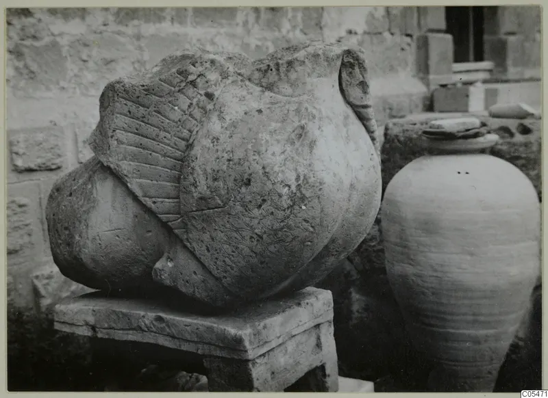 Carved stone sculpture fragment from Soli on a stone block pedestal, with a tall jar beside it.