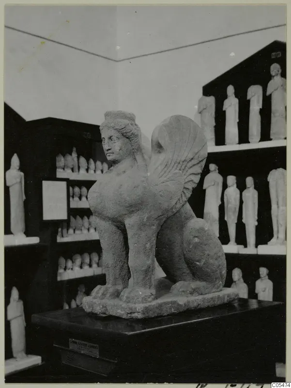 Stone sculpture of a winged creature with a human head and lion-like body, on a dark pedestal in a display room