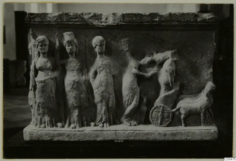 Carved pale stone relief showing four standing female figures and a wheel with an animal at right