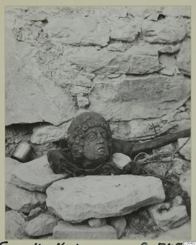 Stone head with curled hair resting among rough stones against a wall, Fasoulla-Kastros