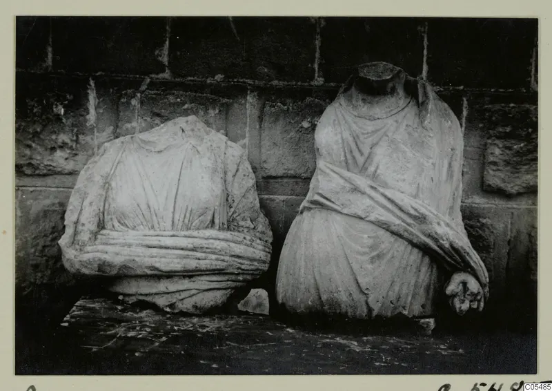 Two headless draped stone statues standing side by side against a stone wall, Arsos