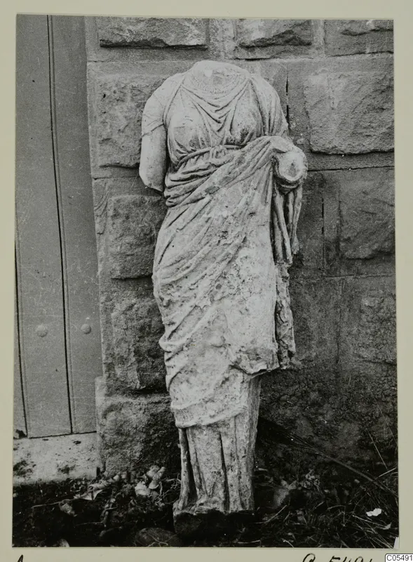 Headless draped stone statue of a standing female figure, photographed against a stone wall beside a wooden door.