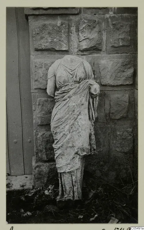 Headless stone statue of a draped female figure standing against a stone wall at Arsos.
