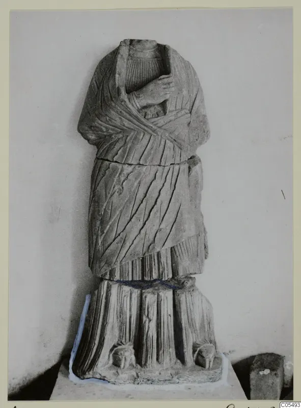 Headless stone statue of a draped standing figure from Arsos, shown against a plain wall.