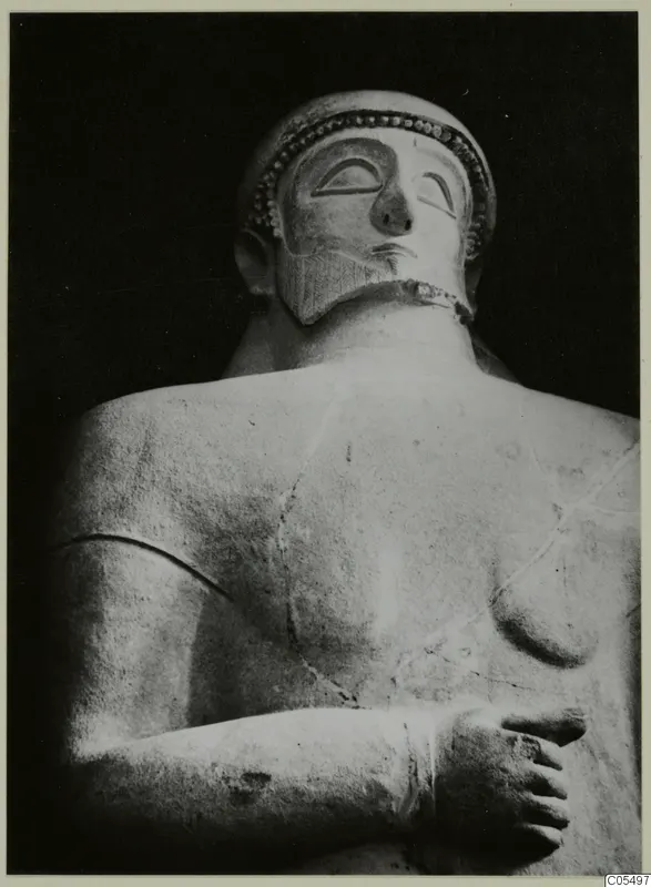 Back and side view of a bearded statue's head, shoulders and folded arm against a dark background.