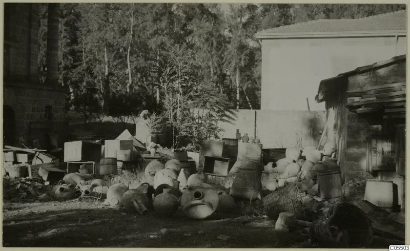 Large pottery jars and vessels piled in a yard beside a stone building and trees, Cyprus Museum