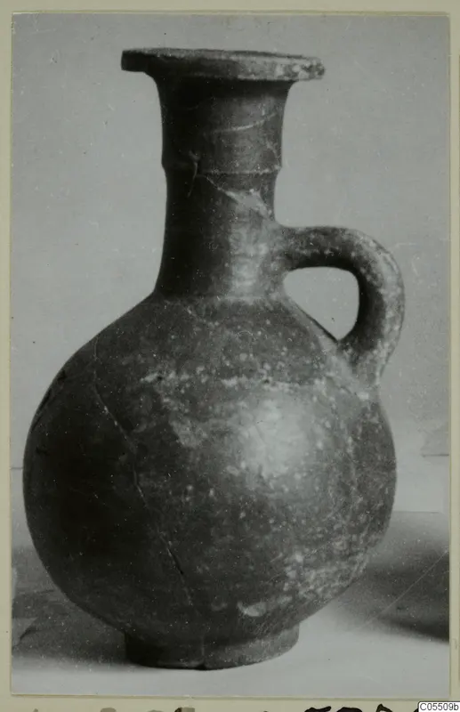 Red-slipped jug with a flared rim, single loop handle and round body, photographed against a grey backdrop.