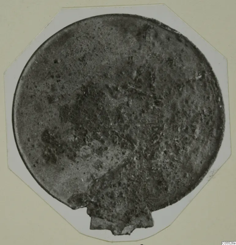 Round bronze plaque from Tomb 25 at Marion, corroded, mounted on a white octagonal card
