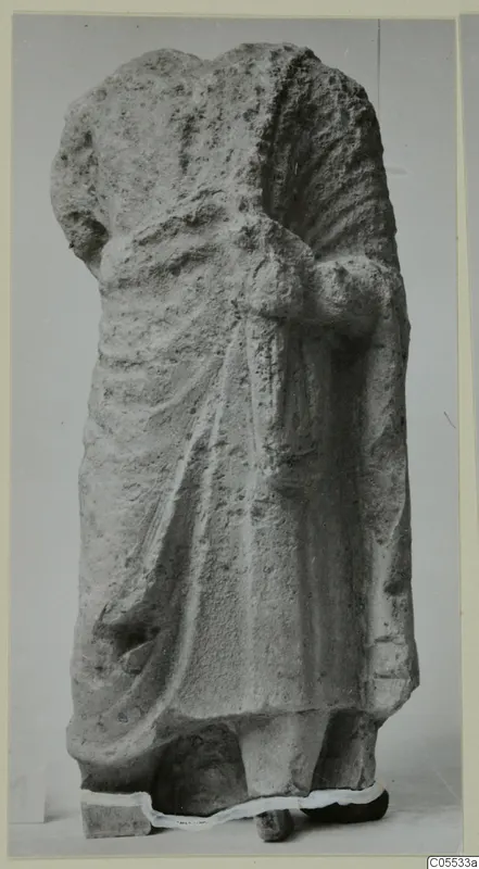 Headless draped standing figure in limestone from Mersinaki, photographed against a grey backdrop.