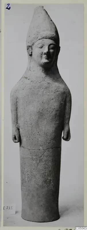 Tall terracotta standing figure with a conical headdress, shown upright against a plain grey background