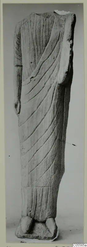 Headless limestone statue of a standing figure in a long draped garment with diagonal folds, on a plinth.