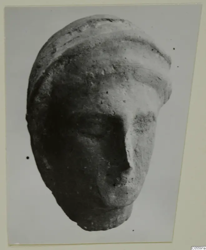 Fragment of a sculpted head from Mersinaki with a banded headdress and broken lower face, shown on a grey backdrop.