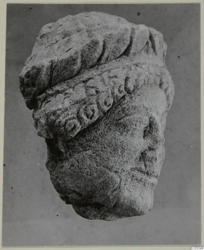 Limestone sculpture of a head wearing a thick wreath-like headdress, from Mersinaki, photographed against a plain grey background.