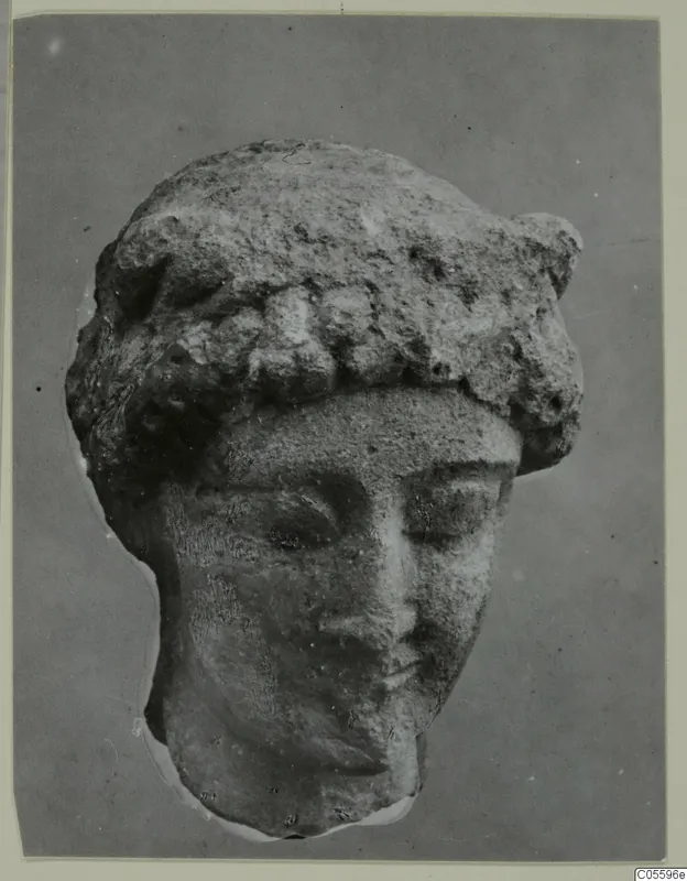 Limestone head of a figure with a rolled headdress and downcast eyes, photographed from the front against a grey backdrop
