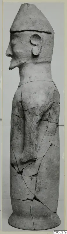 Terracotta standing figure in profile, wearing a tall conical cap, with one arm bent across the body