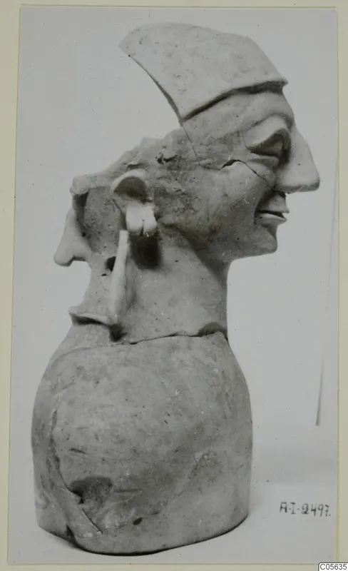Clay bust with two profile faces, one wearing a flat cap-like top, on a rounded base