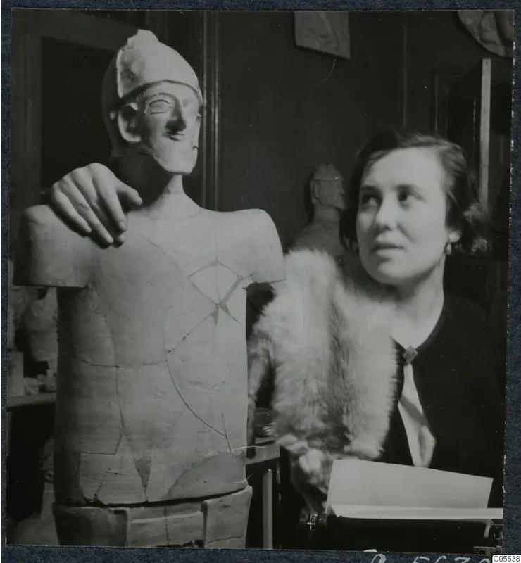 Woman in a fur stole beside a reconstructed statue of a figure with a helmet-like headdress, a hand resting on its shoulder.