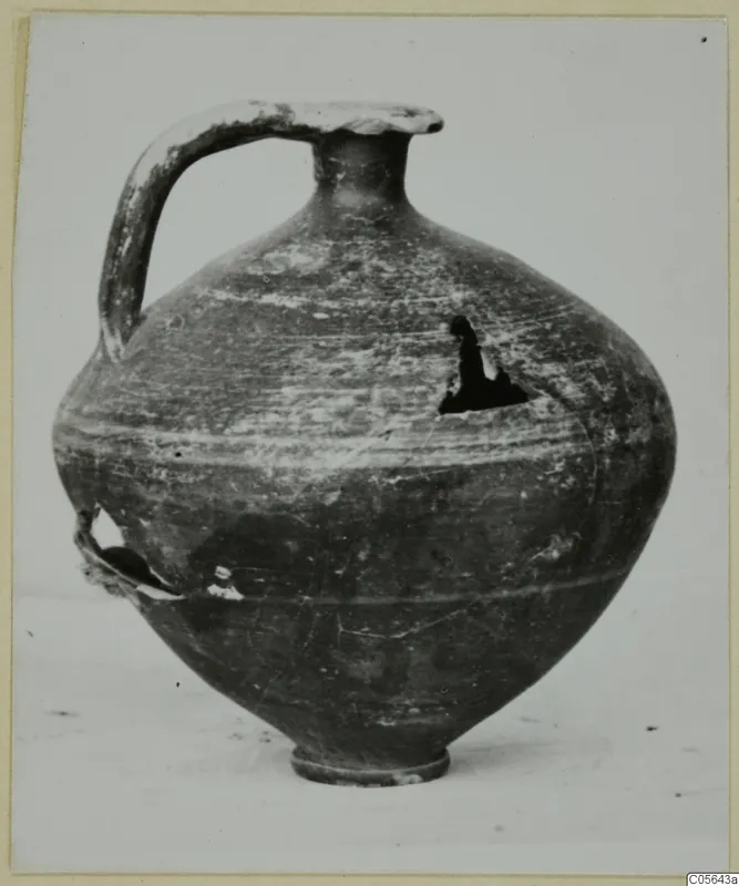 Jug with a tall strap handle, narrow neck and rounded body, with a broken hole in the shoulder and a chipped base
