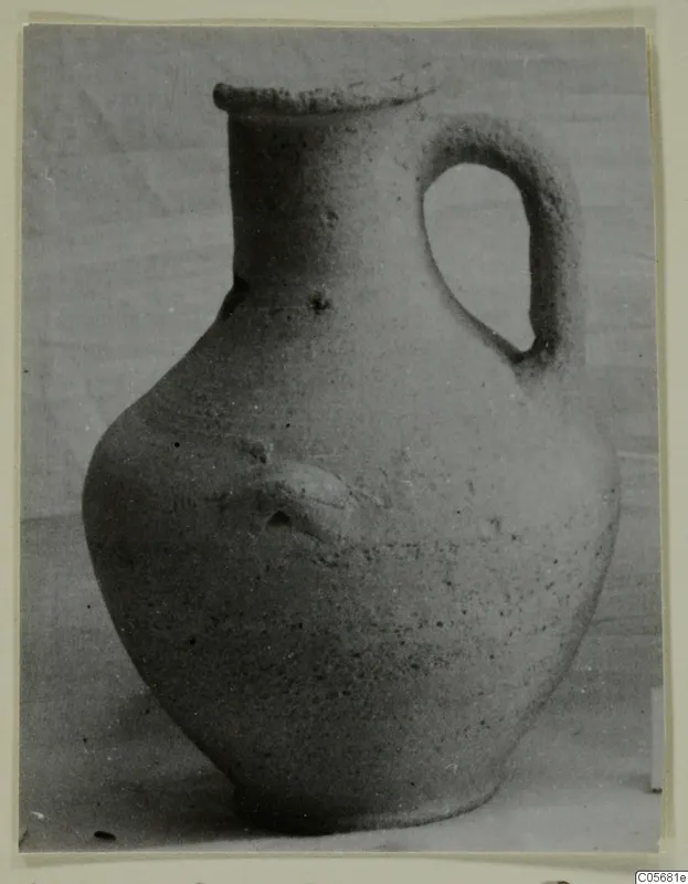 Pottery jug with a single loop handle and a small applied knob on the body, shown against a plain cloth background