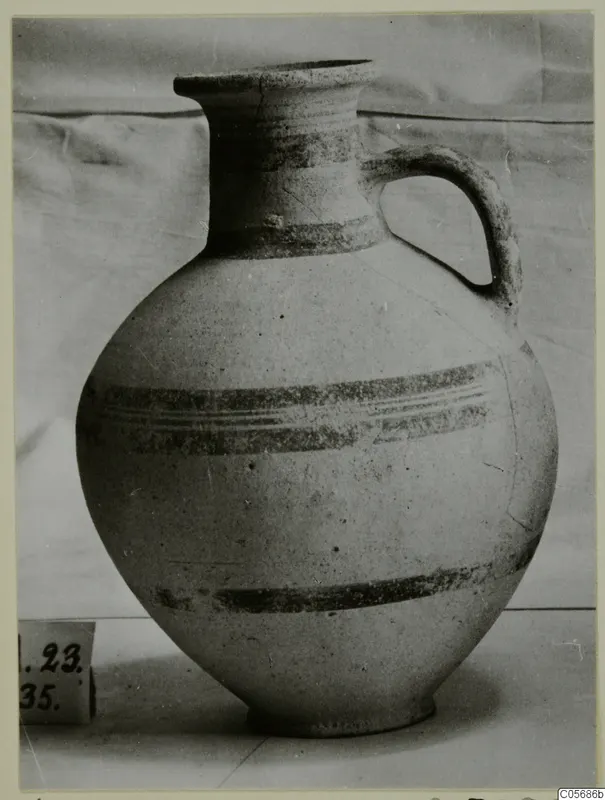 Pottery White Painted IV jug with a single loop handle and dark painted bands, standing on a surface beside a number label.