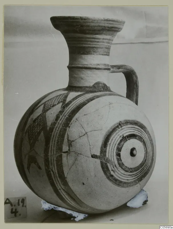 Round-bodied pottery jug with a flaring neck, a handle and painted bands and geometric patterns in dark paint on a pale surface.