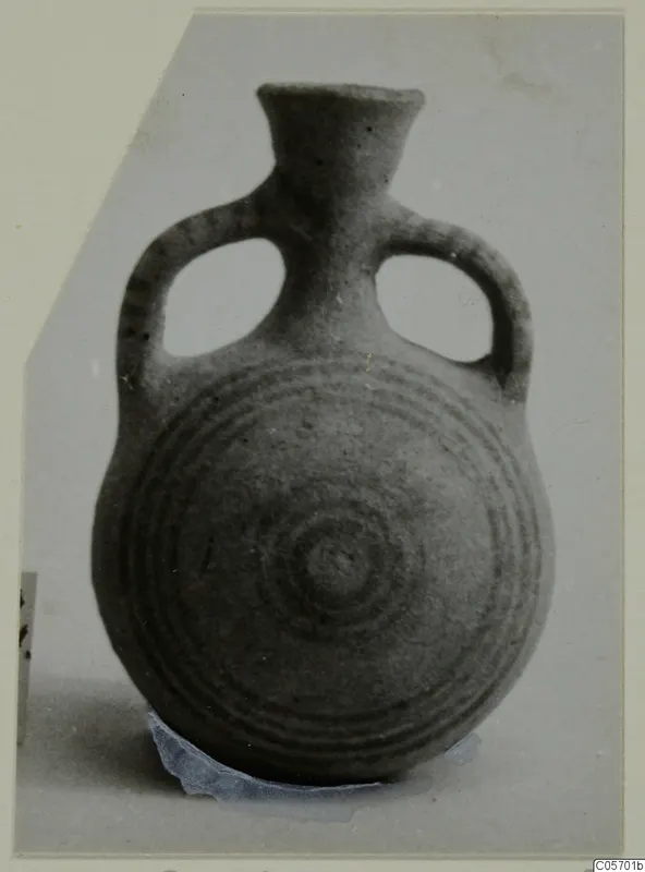 Pottery bottle with two loop handles and concentric circles on the body, photographed on a grey card mount