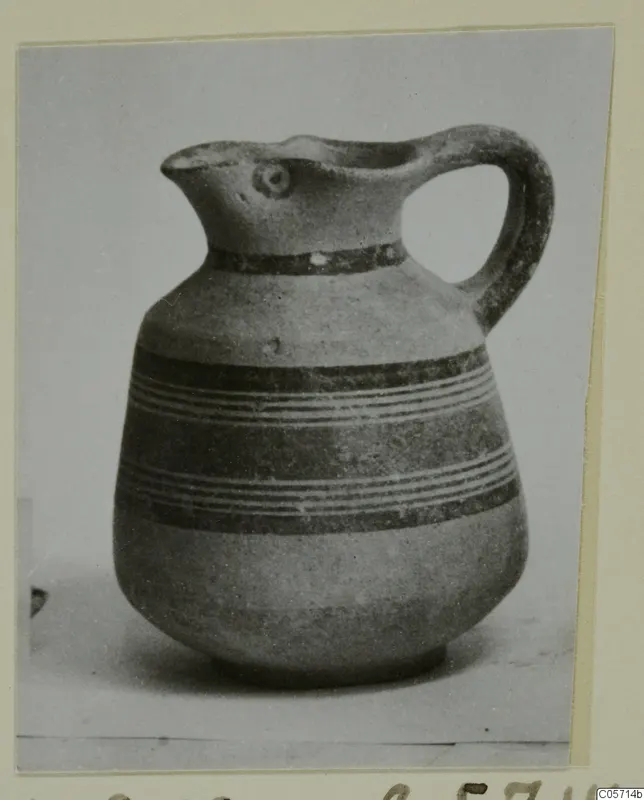 Jug with a pinched spout, a loop handle and dark painted bands, shown in a photograph mounted on a card.