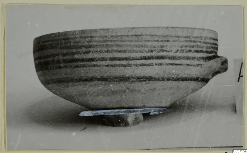 Pottery White Painted IV bowl from Amathus with horizontal dark bands and one side handle, standing on a small stone support.