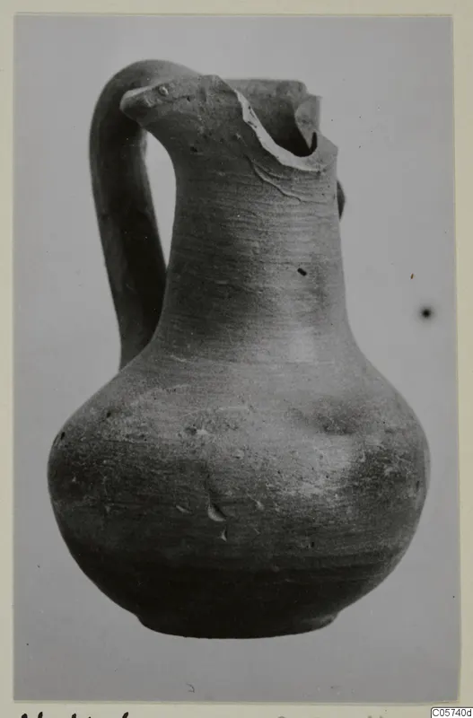 Pottery juglet with a looped handle and a broken rim, photographed against a plain grey background