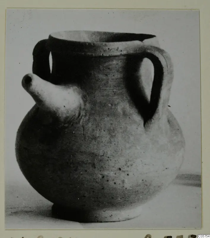 Round pottery vessel with two handles and a short spout, shown on a photo mount with a Cypro-Hell label