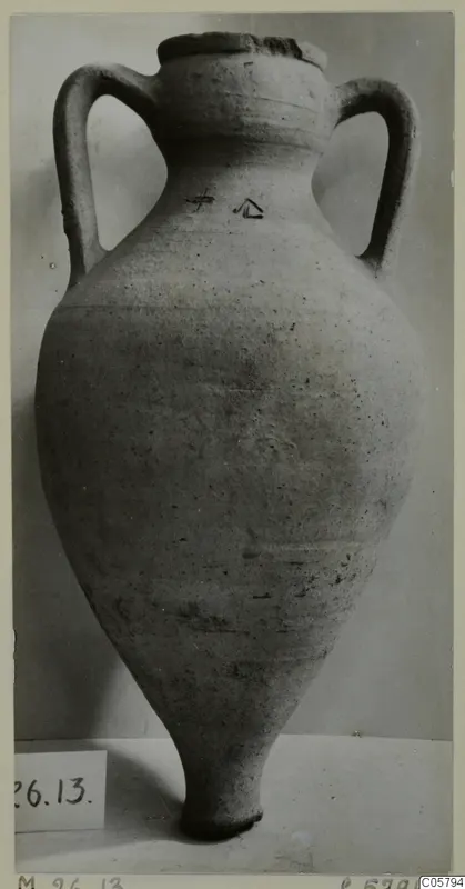 Tall two-handled plain pottery amphora with a chipped rim, standing on a pale surface against a grey backdrop.