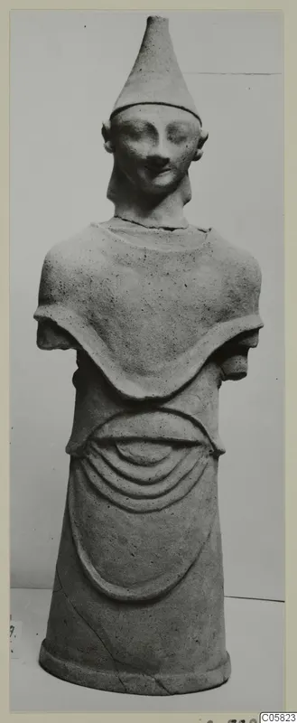 Terracotta figure with a tall conical cap, short stub arms and a pleated skirt, photographed against a grey backdrop