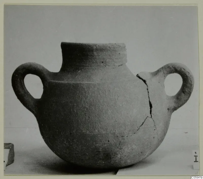 Coarse pottery jug with two loop handles, a tall rim and a cracked body, photographed on a plain surface.