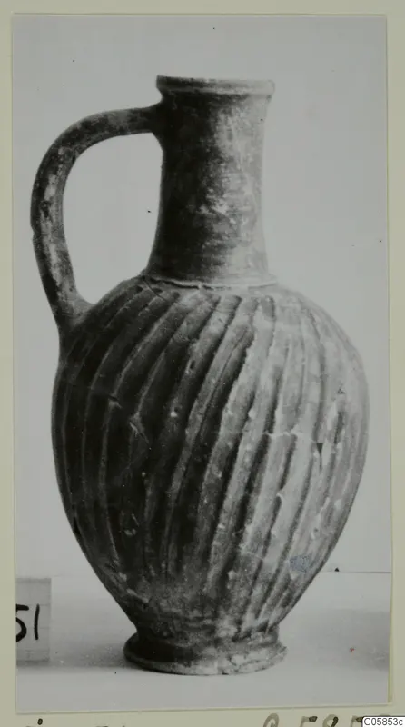 Wheel-made bucchero jug with a single handle and vertically grooved body, photographed on a pale surface
