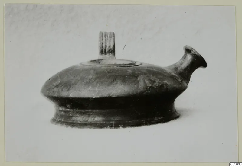 Dark pottery vessel with a spout and a grooved vertical handle on top, photographed on a plain background