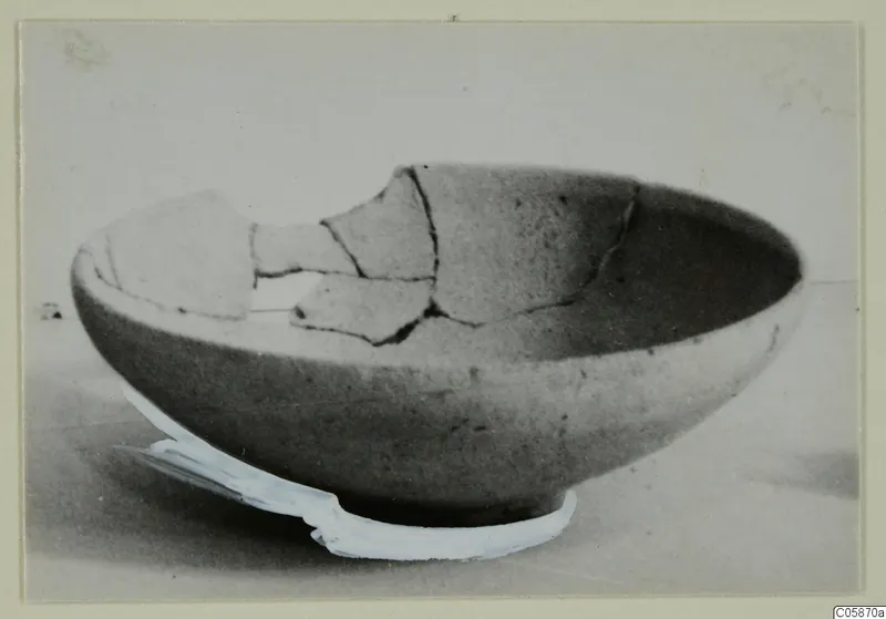 Broken pottery bowl with a rounded body and wide mouth, photographed on a pale surface with a white strip beneath.