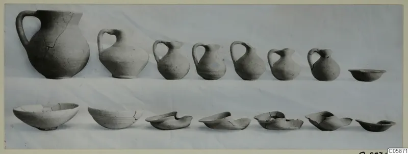 Row of handled jugs and a row of bowls, pottery from Tomb 1 at Idalion, laid out on a pale cloth.