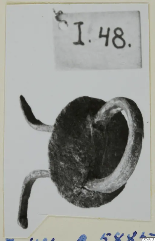 Corroded metal mounting with a loop and two bent prongs, photographed on a card with a number label