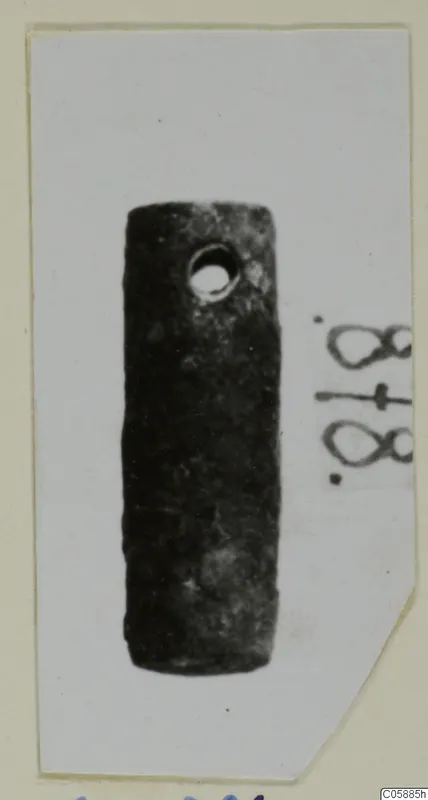 Cylindrical dark metal fragment with a round hole near one end, photographed on a mounted card.
