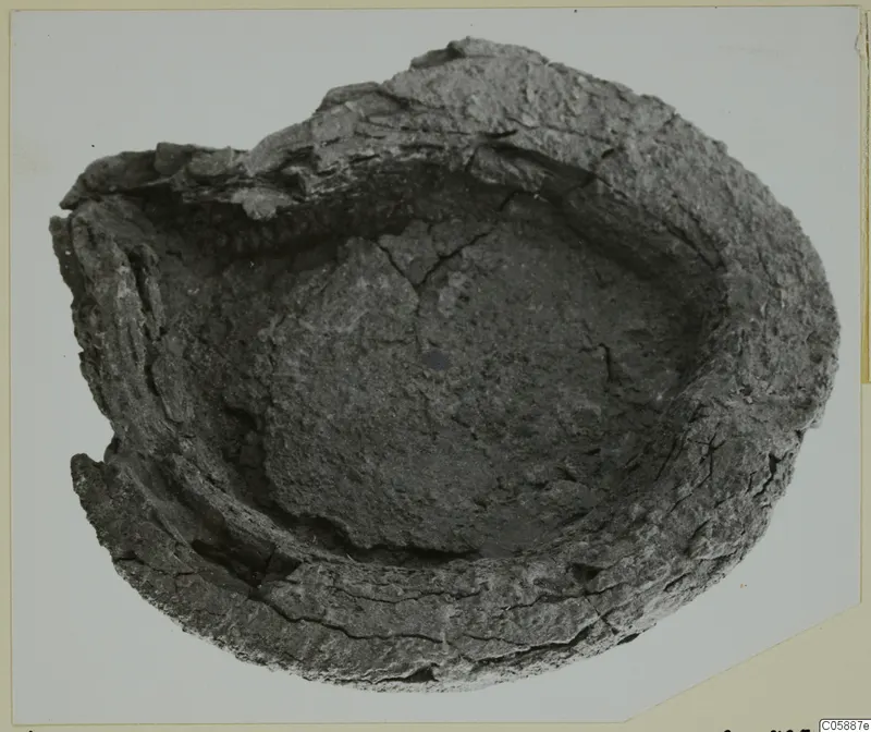 Heavily corroded metal object with a shallow bowl form and broken rim, photographed from above on a pale card.