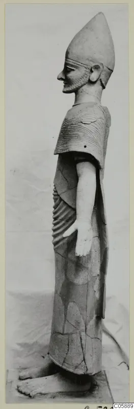 Side view of a standing terracotta figure with a tall conical headdress, a short beard and a ribbed cape.