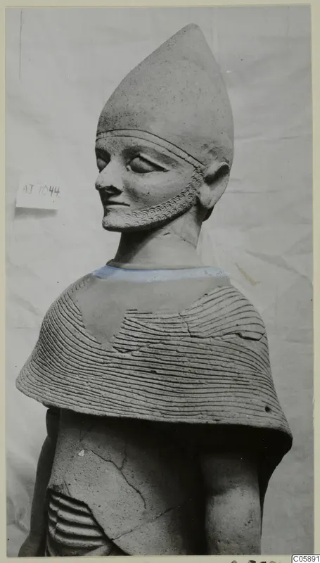 Terracotta statue with a tall conical head covering, braided beard and cape with horizontal incised lines