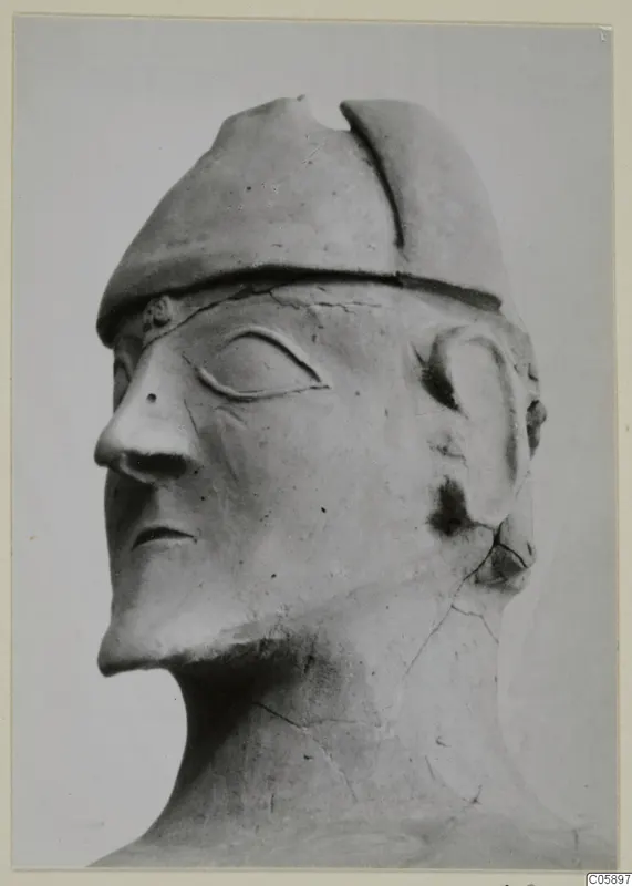 Terracotta head with a cap-like headdress and a long nose, shown in close view against a plain grey backdrop.