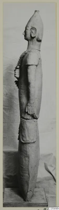 Side view of a tall terracotta statue of a standing figure with a tall head, on a wooden base