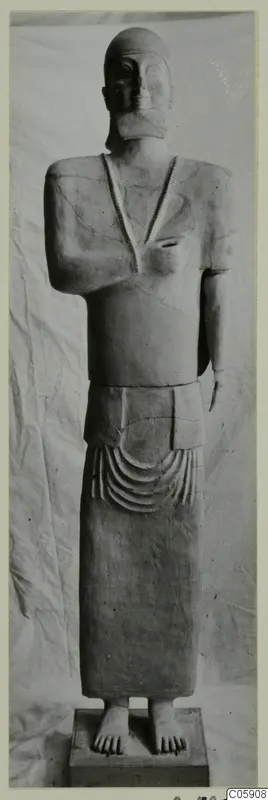Standing terracotta statue of a figure with a cap and necklace, on a rectangular base, shown against a draped cloth