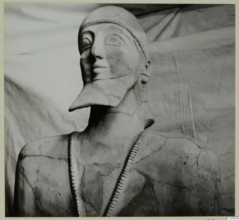 Large terracotta statue of a figure with a long pointed beard and a cap, seen from the chest up against a cloth backdrop.