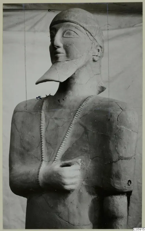 Terracotta Neo-Cypriote statue of a standing figure with a pointed beard and cap, one hand held against the chest.